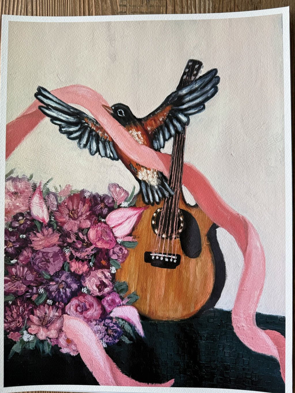 A rectangular art print  depicting a robin bird with spread wings resting on a wooden guitar, wrapped in a pink ribbon, with a bouquet of pink and purple flowers at the lower left, shown against a light background with a white border.