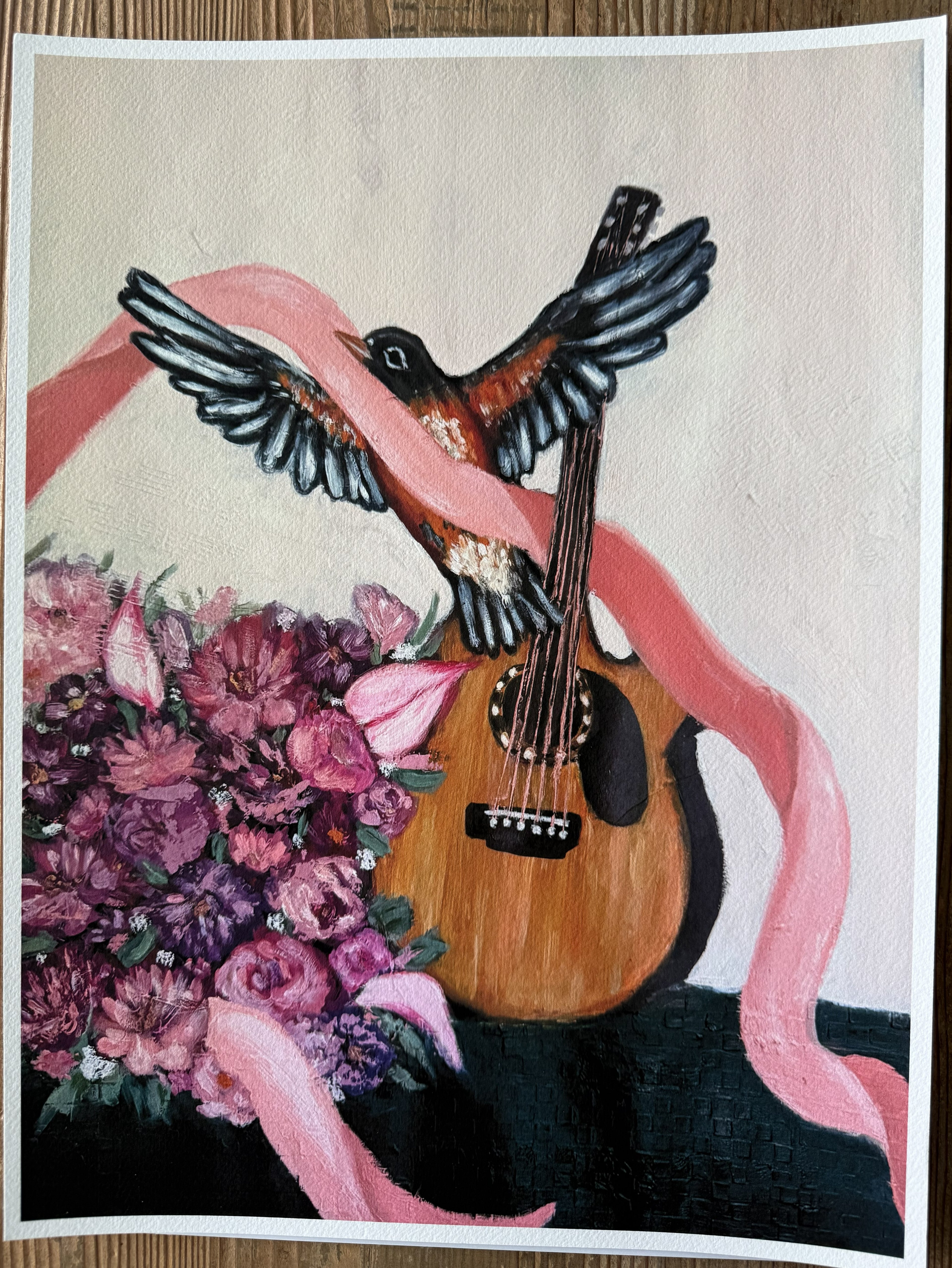 A rectangular art print  featuring a bird with spread wings perched on a wooden guitar, wrapped in a pink ribbon, with a bouquet of pink and purple flowers at the lower left, shown against a light background.