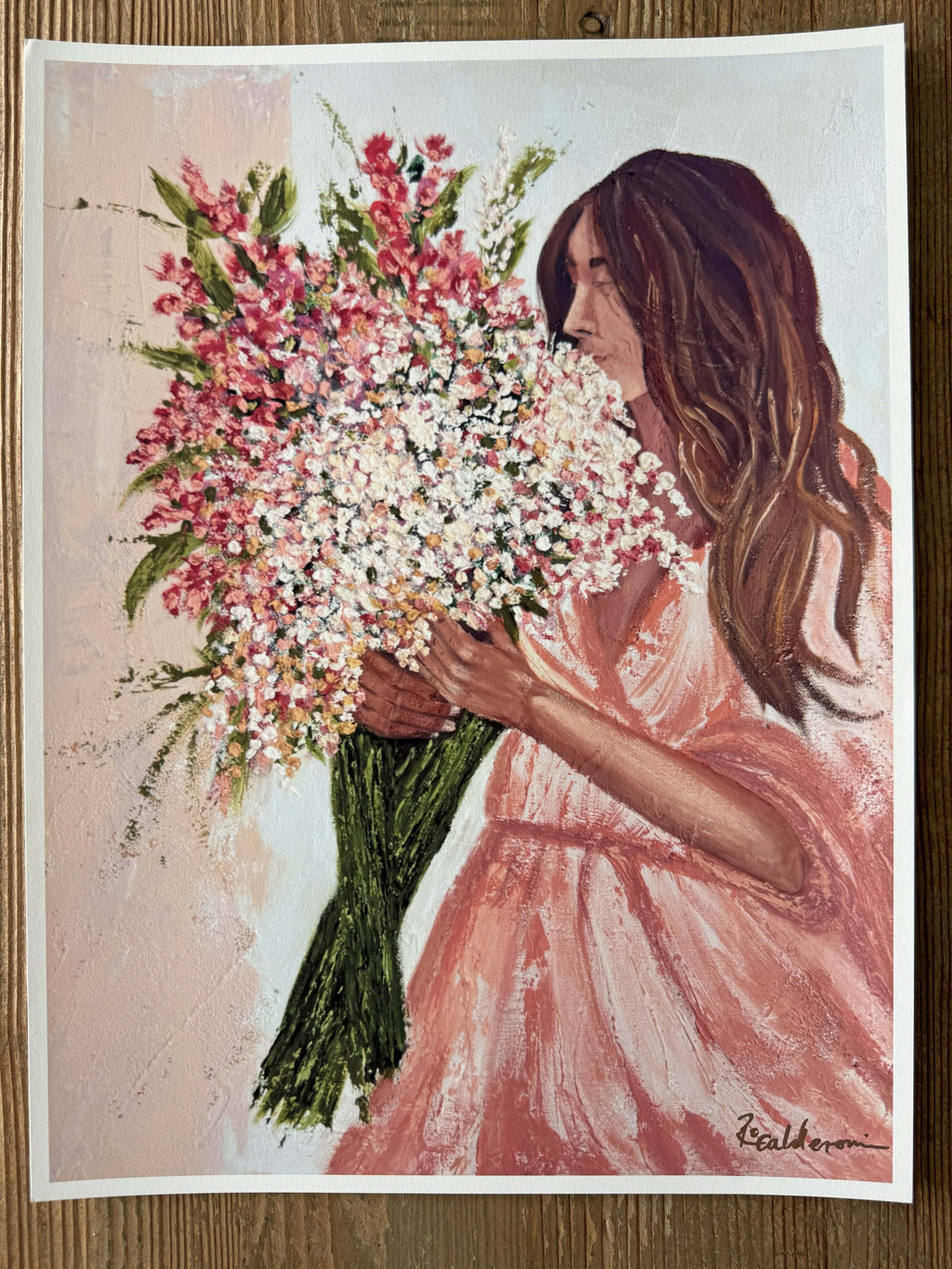 A rectangular art print featuring a painted woman with long brown hair holding a large bouquet of pink, white, and green flowers, shown against a light background with a white border. The print is photographed flat on a wooden surface.