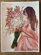 A rectangular art print featuring a painted woman with long brown hair holding a large bouquet of pink, white, and green flowers, shown against a light background with a white border. The print is photographed flat on a wooden surface.