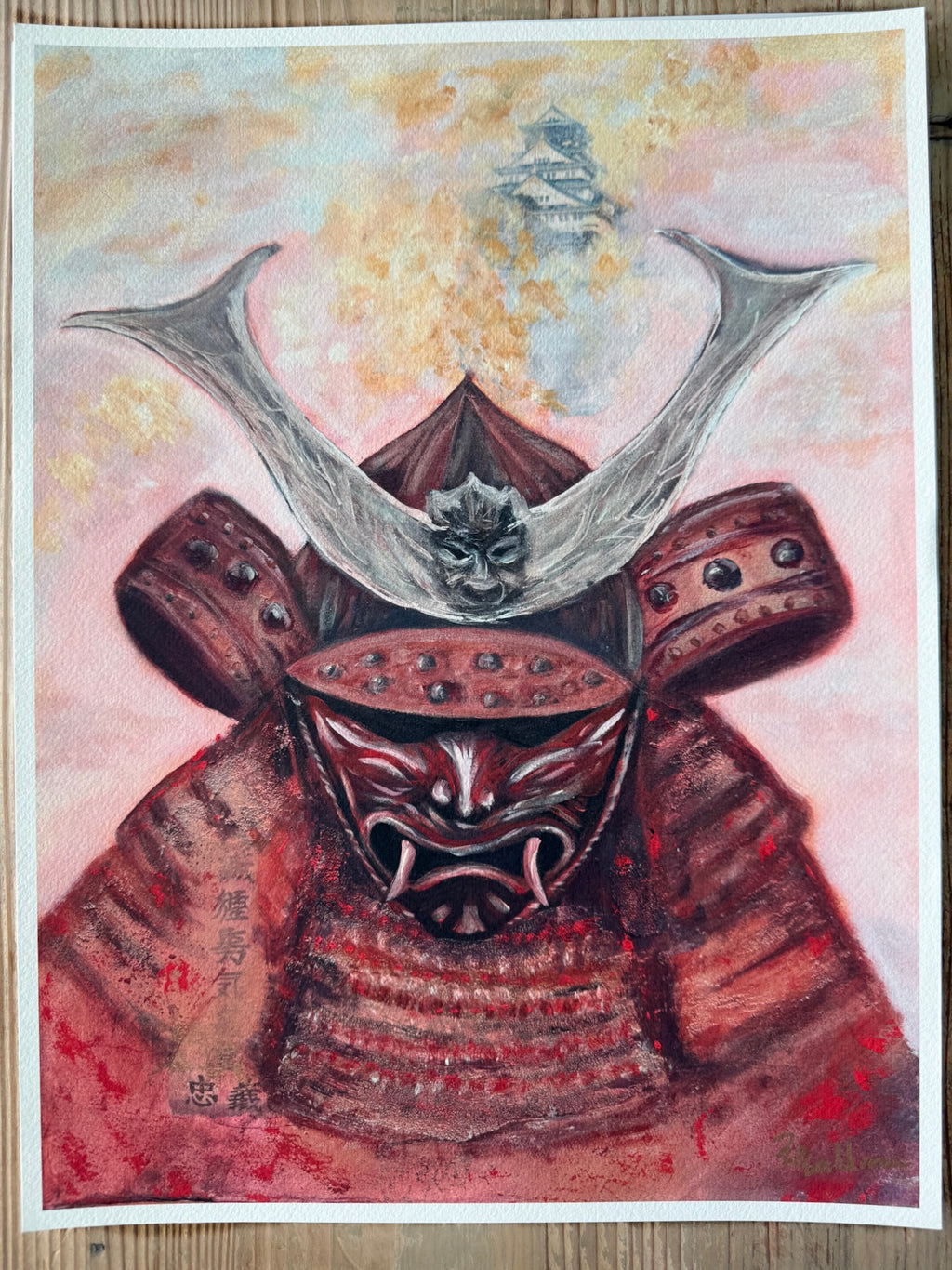 A painted artwork of a samurai warrior in red armor with a large horned helmet, shown on a white paper sheet against a light wood background.